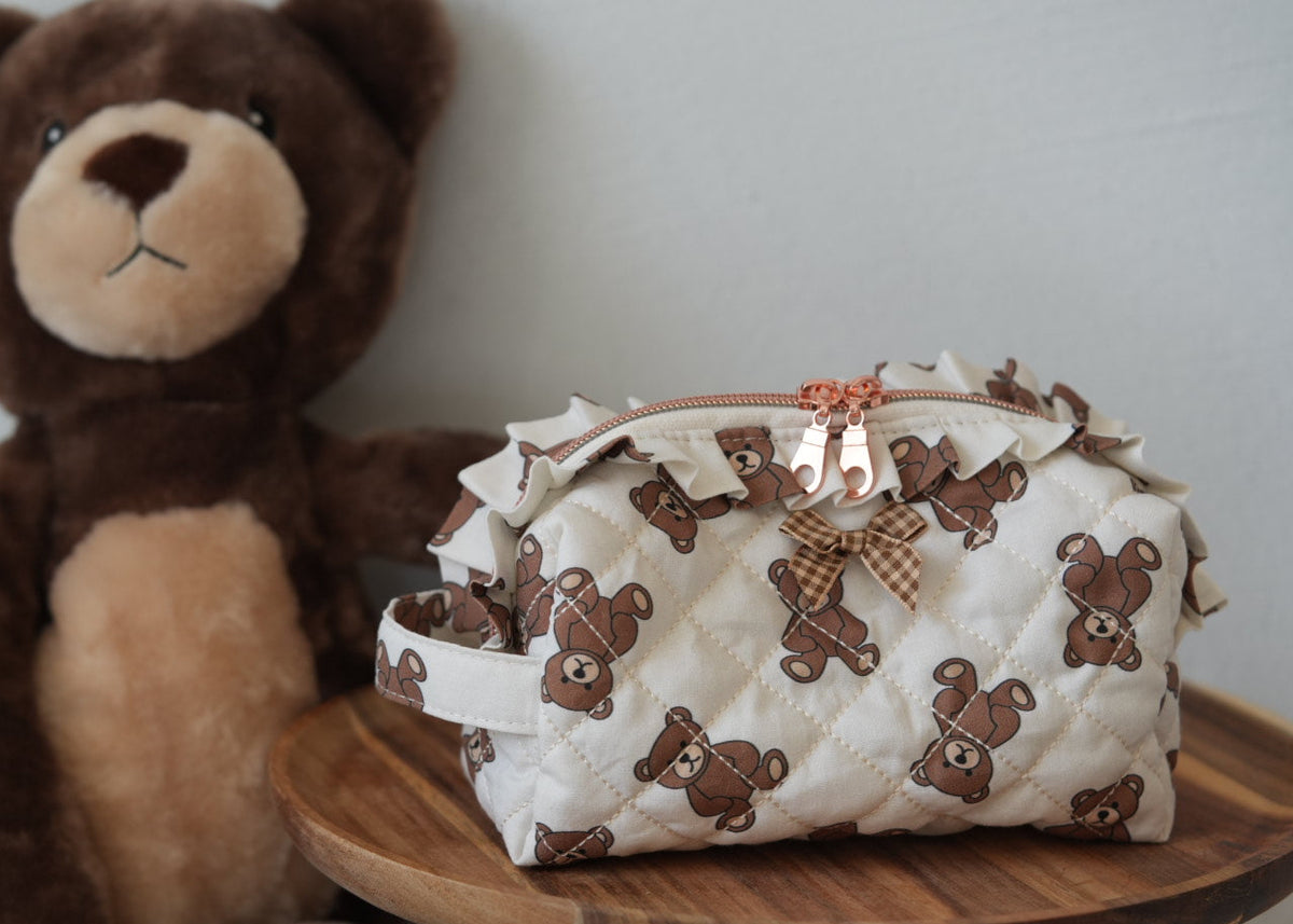 A quilted cream-colored cosmetic pouch with a brown teddy bear print, featuring a ruffled trim and a small bow, displayed on a wooden stool next to a large plush teddy bear.