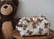 A quilted cream-colored cosmetic pouch with a brown teddy bear print, featuring a ruffled trim and a small bow, displayed on a wooden stool next to a large plush teddy bear.