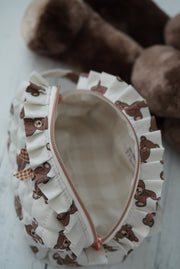 A close-up of a ruffled fabric zip pouch with a brown teddy bear print, shown open with a cream plaid lining and a rose gold zipper. A plush brown teddy bear toy is blurred in the background on a light surface.